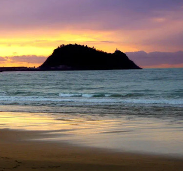 Atardecer en la playa de Getaria con colores cálidos reflejados en el agua y el icónico monte San Antón al fondo.